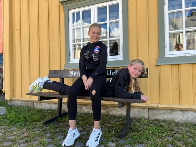 En ung jente sitter oppå ryggen til en annen jente som ligger på en benk. De har idrettsklær på seg.